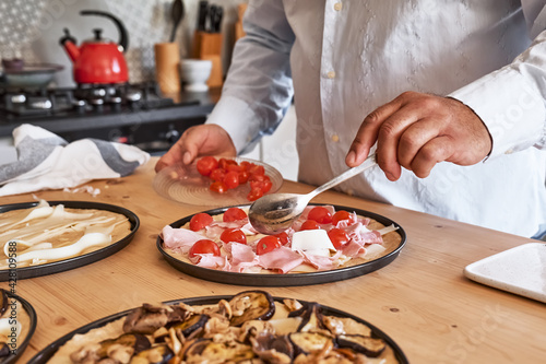 Fototapeta Naklejka Na Ścianę i Meble -  Homemade Italian pizza. Man in kitchen putting ingredients on pizza like tomato, mushrooms, eggplant, cheese and ham. Selective focus.