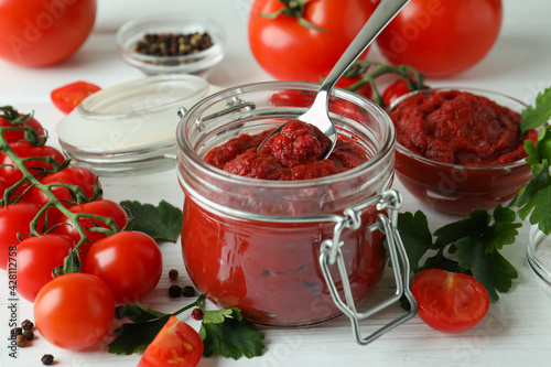 Jar and spoon with tomato paste on white wooden table with ingredients