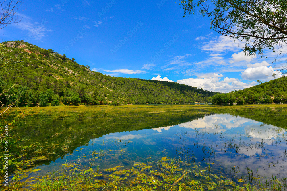 Vue du lac de Sainte Suzanne (dit lac de Carces) avec les reflets des montagne dans l'eau et les plantes aquatiques au premier plan sou un cliel bleu azur parsemé de nuages blancs