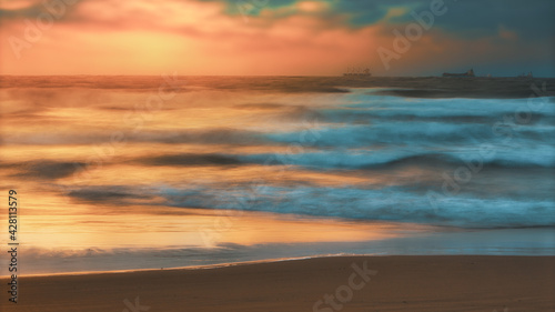 sunset on the beach with waves in front and ships far in the background © syanai