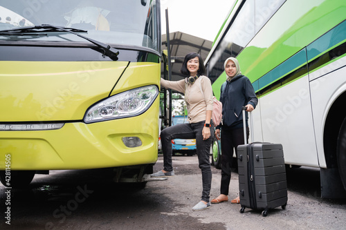 a short-haired woman with headphones and a bag smiling at the camera as she steps up to the bus door with a woman in a headscarf carrying a suitcase behind her while going by bus