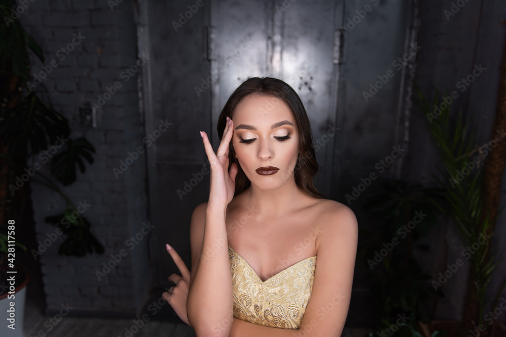 Fototapeta premium Close up beauty portrait of girl, holds her hand to head, headache. Brunette with brown eyes, professional evening make-up, dark lipstick, smoky eyes in gold corset. Festive, party. Silver background