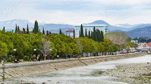 Beautiful embankment of Adler on bank mountain river Mzymta against background of peaks Caucasus Mountains. Famous resort in   Sochi in south of Russia in spring. Adler, Russia - March 16, 2021 photo