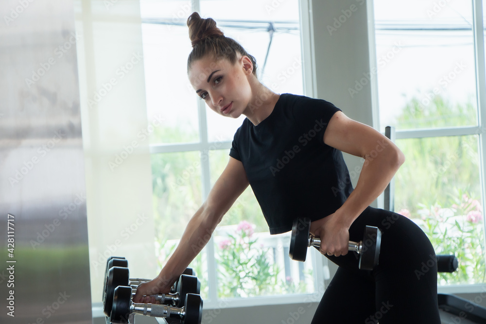 Fototapeta premium A beautiful caucasian white woman holding a body dumbbell standing in front of a mirror at a fitness gym.