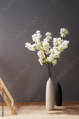 Still life: flowering branches of white sakura in white vase near wooden ladder and empty black vase on the wooden floor. The splendid bouquet is outlined against gray wall. 