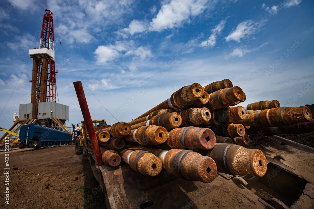 Stock of rusted oil drill pipes on foreground, in focus. Drill rig (left) is blurred. Blue sky, clouds. Uralsk region, Kazakhstan. Oil deposit Zhaikmunai. Stock of rusted oil drill pipes on foreground, in focus. Drill rig (left) is blurred. Blue sky, clouds. Uralsk region, Kazakhstan. Oil deposit Zhaikmunai.