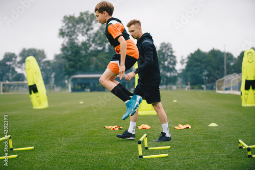 Young man jumping over practice hurdles in soccer training. Soccer coach watching boy on training. Player of youth football academy with coach on pitch © matimix
