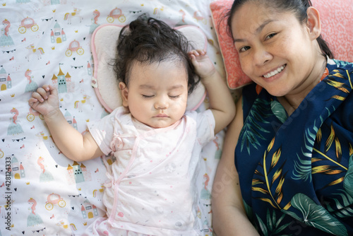 Cute little Asian baby girl sleeping on bed beside mother, mother smile and looking at camera