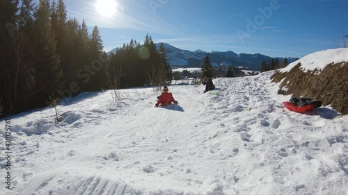 Kid in red jacket jump in slow motion on sled while sledding down from snowy hill in winter during sunny day in Zakopane.