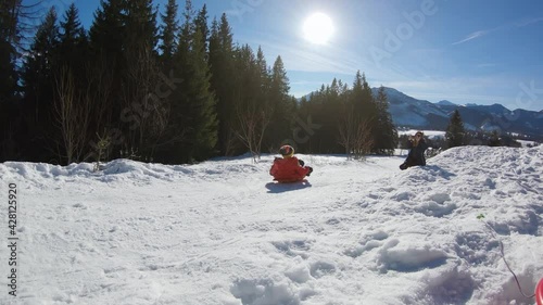 Kid in red jacket jump in slow motion on sled while sledding down from snowy hill in winter during sunny day in Zakopane.