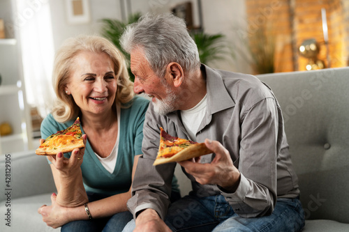 Cheerful husband and wife sitting on sofa at home. Happy senior woman and man eating pizza while watching a movie.