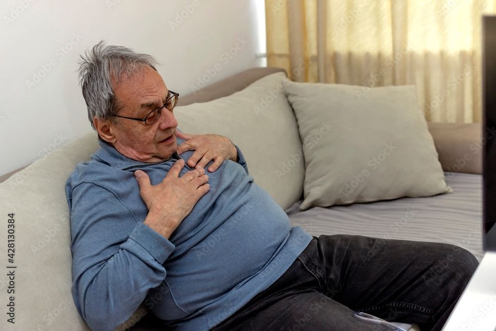 Shot of an uncomfortable looking old man holding his chest in discomfort while being seated on a couch at home. Overweight senior man holding breast because of heart infarction. Health care concept. Shot of an uncomfortable looking old man holding his chest in discomfort while being seated on a couch at home. Overweight senior man holding breast because of heart infarction. Health care concept.