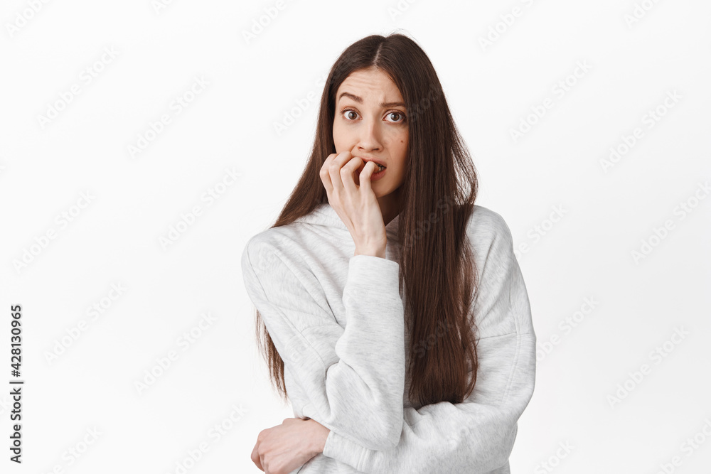 Worried young woman biting fingers and looking at camera, have anxiety, bite fingernails nervous, standing in hoodie against white background Worried young woman biting fingers and looking at camera, have anxiety, bite fingernails nervous, standing in hoodie against white background