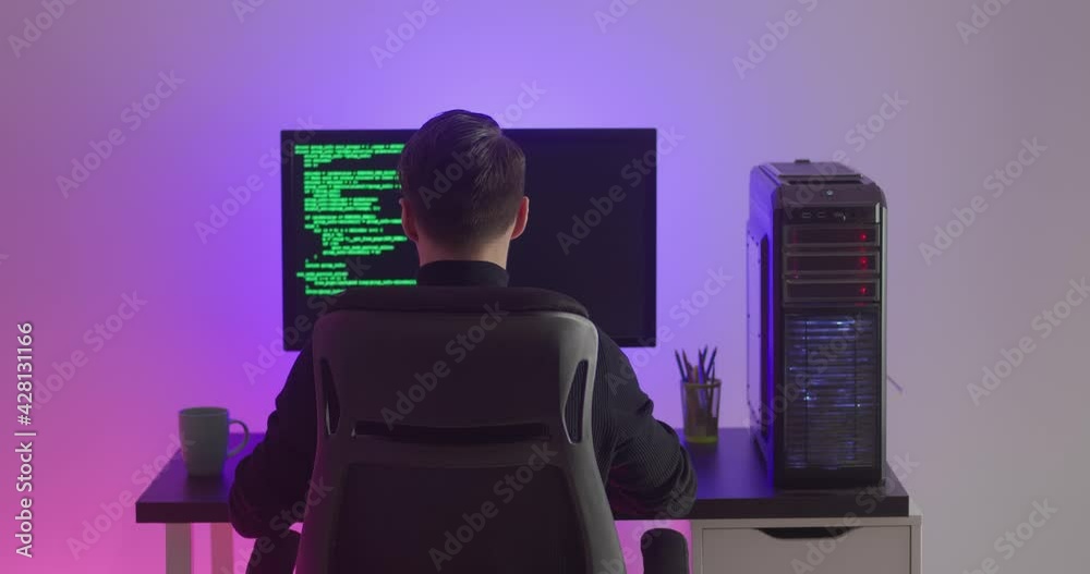 Rear view of man programmer coding network security software indoors in neon lights at night. Hacker typing code on pc computer screen in dark office Rear view of man programmer coding network security software indoors in neon lights at night. Hacker typing code on pc computer screen in dark office