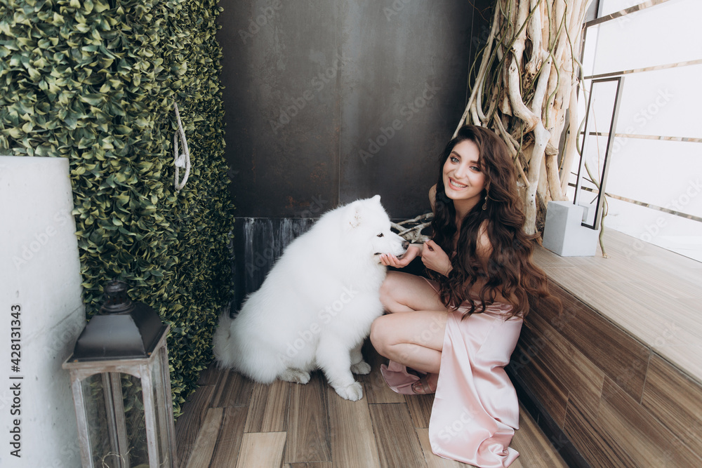 Beautiful, stylish, and elegant girl brunette, model, with makeup and curly hair in a peach-colored silk dress with a white dog of breed malamute for a magazine photoshoot in fashion style. Beautiful, stylish, and elegant girl brunette, model, with makeup and curly hair in a peach-colored silk dress with a white dog of breed malamute for a magazine photoshoot in fashion style.