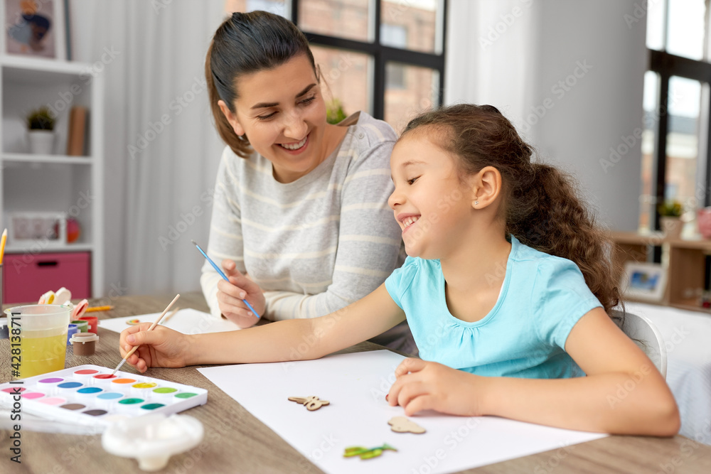 family, motherhood and leisure concept - happy smiling mother spending time with her little daughter drawing or painting wooden chipboard items with colors at home