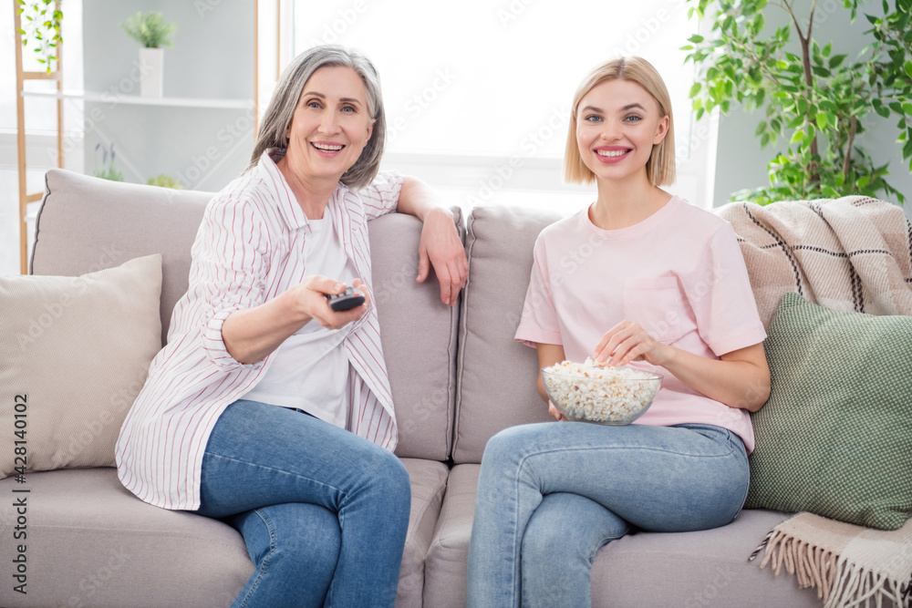 Photo of happy grey haired old lady and blonde woman sit sofa watch tv eat popcorn indoors inside house home
