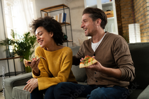 Cheerful young couple sitting on sofa at home. Happy woman and man eating pizza while watching a movie.