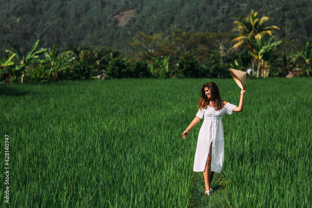 A beautiful woman walks on rice terraces on the island of Bali. A beautiful woman in a white dress and a conical hat walks through green rice fields. Tourist traveling in Indonesia. Copy space