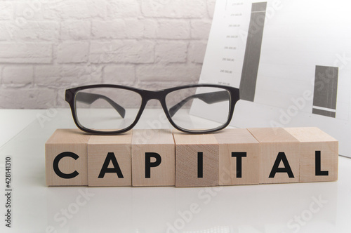 The word CAPITAL is written on wooden cubes standing on the white surface of the table, the GLASSES are on the cubes