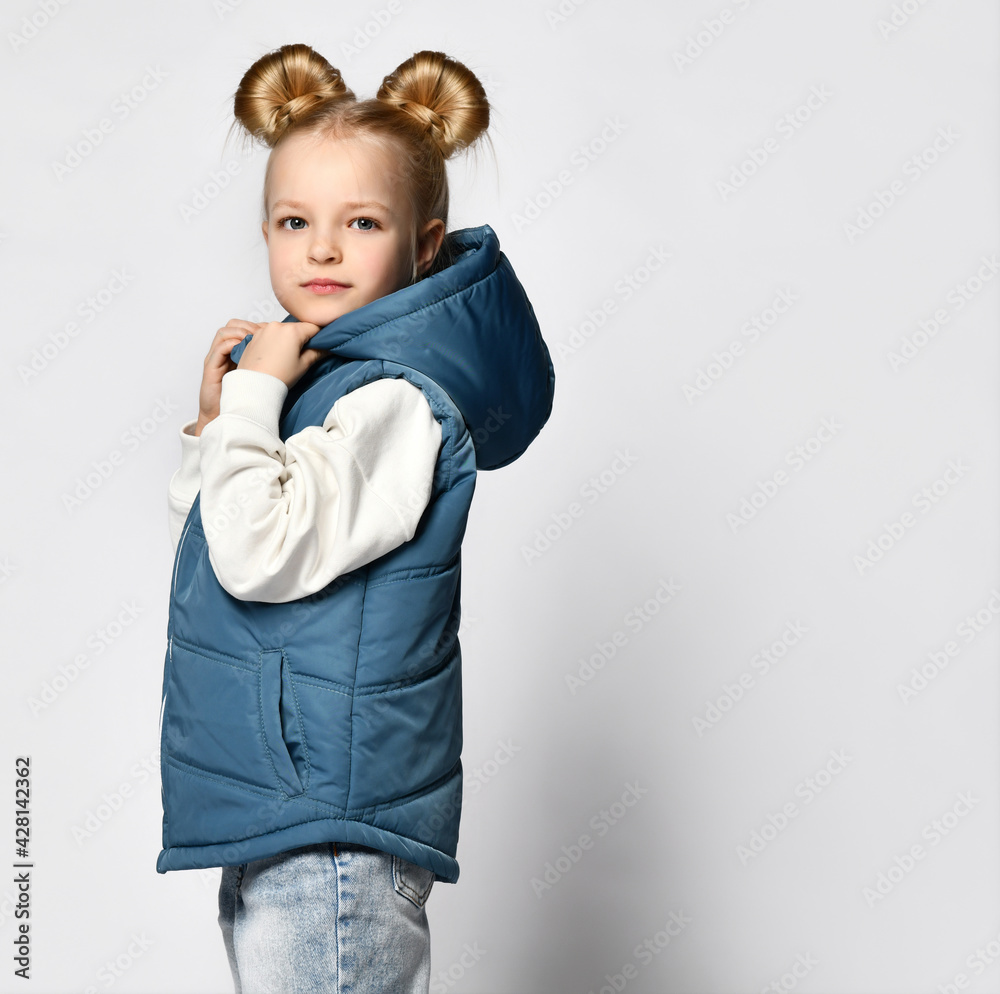 Side view of a little girl in warm outerwear blue sleeveless vest, jeans and a sweatshirt, posing for the camera isolated on white background. Children's fashion clothing and fashion advertising Side view of a little girl in warm outerwear blue sleeveless vest, jeans and a sweatshirt, posing for the camera isolated on white background. Children's fashion clothing and fashion advertising