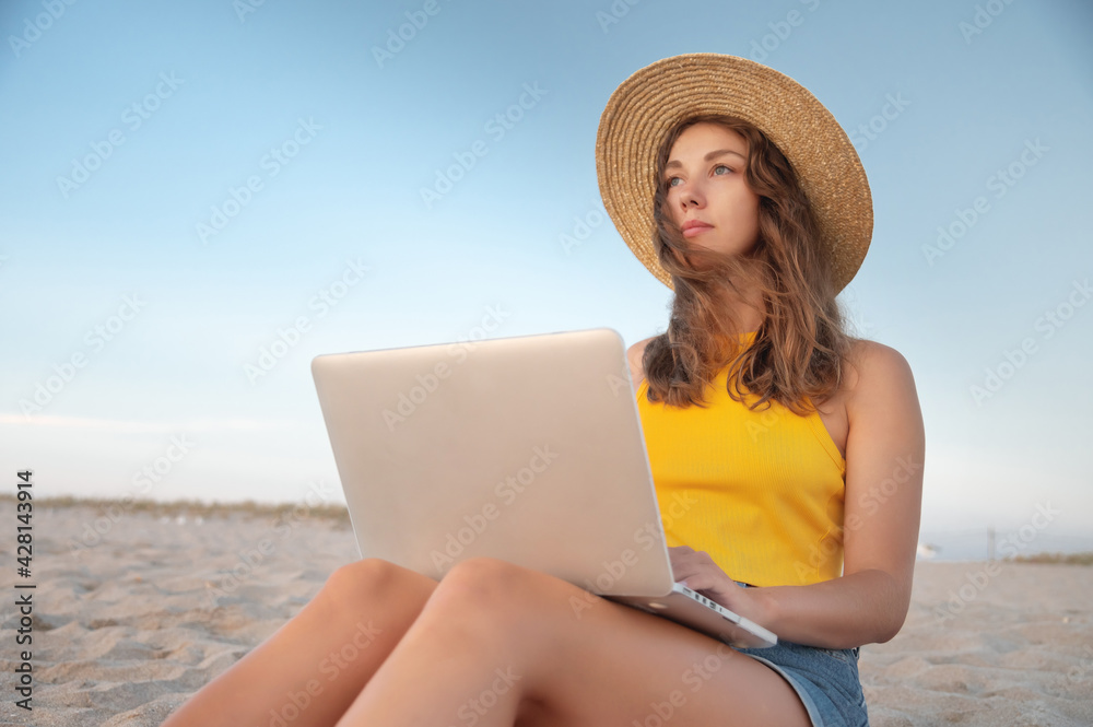 young woman freelancer, in a hat, sits on a sandy beach by the sea with a laptop on her lap