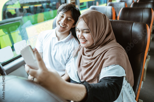 Asian Muslim couples smile at their cellphone camera while taking a selfie together while sitting on the bus