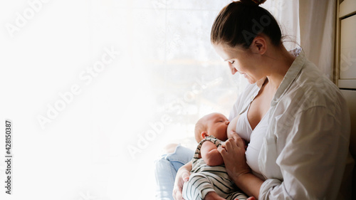 Cute caucasian mom and newborn baby, mother breastfeeding baby by the bed in a bright white bedroom. Top view, toning and lifestyle in a real interior photo