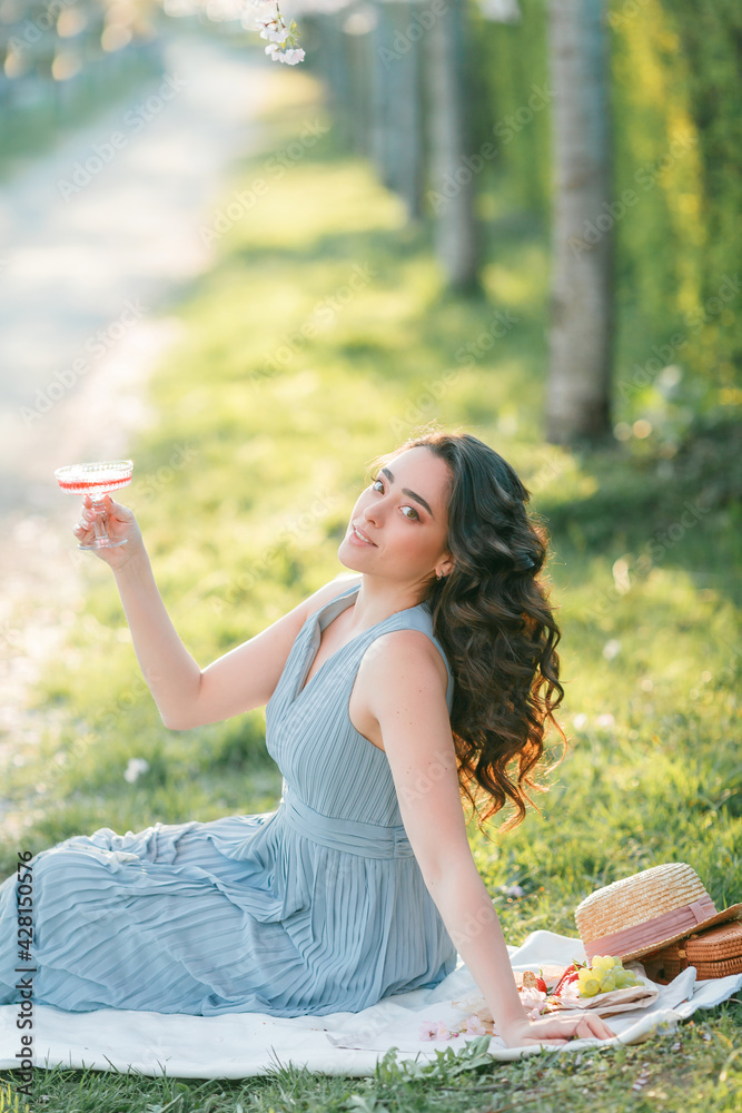 Naklejka premium Beautiful young woman with curly hair drinks pink champagne in the blooming sakura garden. Picnic in nature.