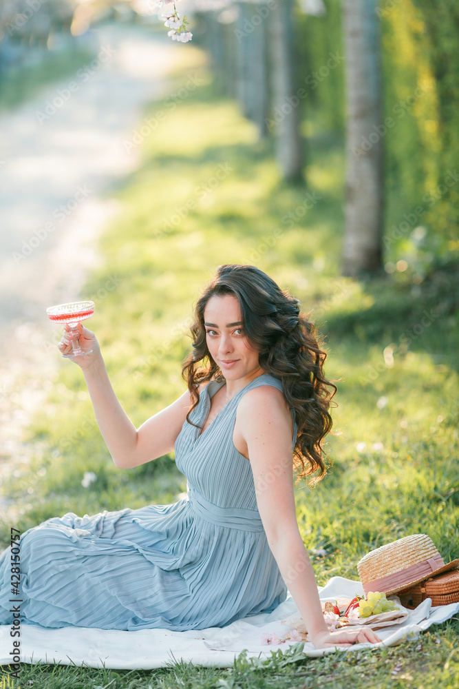 Naklejka premium Beautiful young woman with curly hair drinks pink champagne in the blooming sakura garden. Picnic in nature.