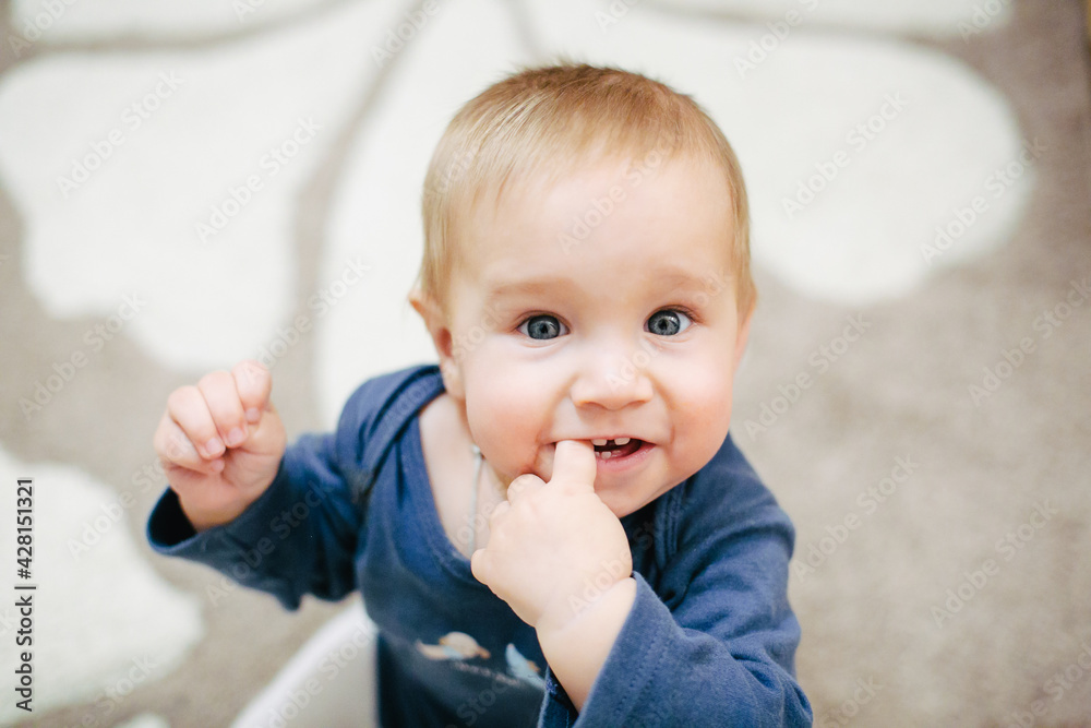 Close-up portrait of child in blue shirt smiling and attentively looking at camera and having his finger in his mouth. Blurred beige background Close-up portrait of child in blue shirt smiling and attentively looking at camera and having his finger in his mouth. Blurred beige background