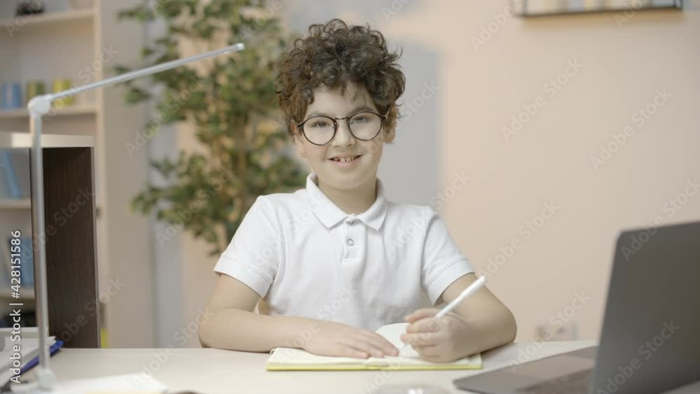 Smart little boy in eyeglasses writing in notebook, smiling at camera, education Smart little boy in eyeglasses writing in notebook, smiling at camera, education