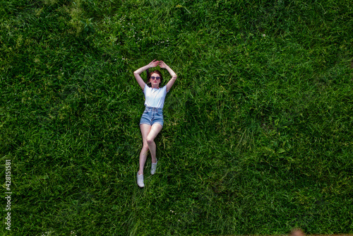 Portrait of happy cute young woman with short hair in casual black and white striped shirt, red round eyeglasses lying down on green grass and looking at camera through glasses.outdoor summertime shot