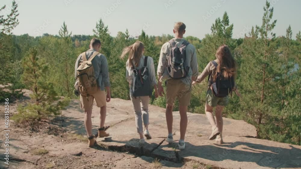 Slowmo tracking shot of happy young women and men with backpacks walking to edge of cliff and enjoying scenery. They are raising arms and cheering Slowmo tracking shot of happy young women and men with backpacks walking to edge of cliff and enjoying scenery. They are raising arms and cheering
