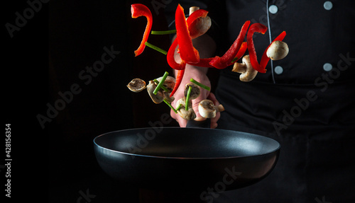 Hand of a professional chef tosses pieces of vegetables and mushrooms on a frying pan on a black background. Restaurant cooking concept. Free advertising space