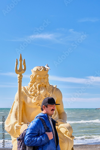 A middle-aged Asian man with a backpack walks along the seaside promenade past a sculpture of Neptune. © IrinaK