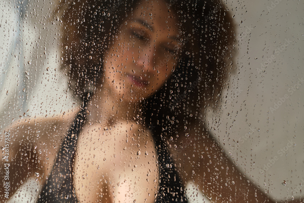 Sensual portrait of young woman taking a shower. Defocused female looks through the glass of the shower stall. Art portrait. Feminine beauty. Skincare wellness.