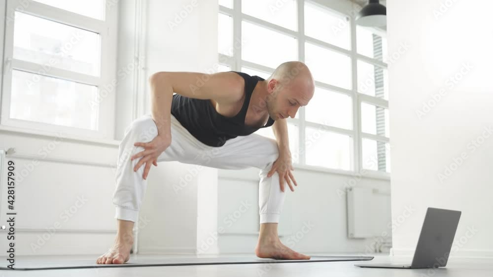 Young man working out, online training via webcam, using laptop, going to perform sumo squat exercises, revolved temple, goddess, sumo wrestler pose.