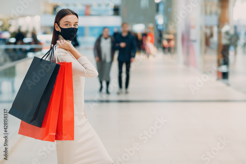 Young adult woman in protective medical face mask carrying paper shopping bags in hands at shopping mall