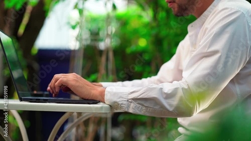 Young asian man with mustaches and a beard, is sitting at the table in garden, is typing on laptop. Working outside.