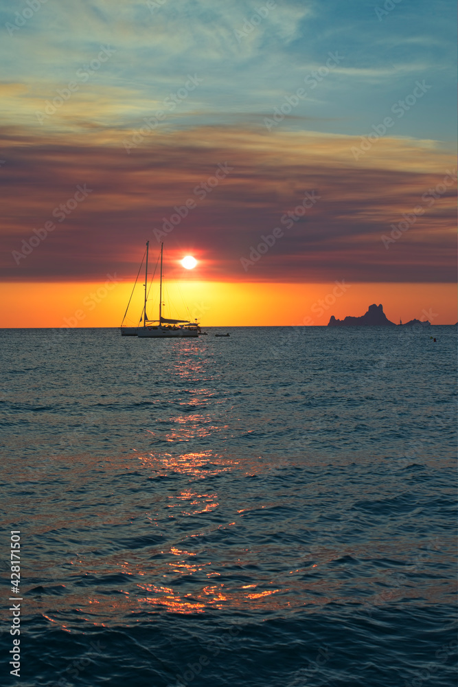 Two sailboats at anchor at sunset in front of a beach on the island of Formentera in Spain. In the background the sun on the horizon and the small island of Es Vedra. Two sailboats at anchor at sunset in front of a beach on the island of Formentera in Spain. In the background the sun on the horizon and the small island of Es Vedra.