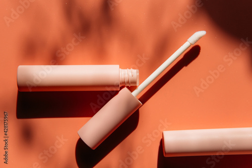 open tube with a brush for liquid lip gloss, liquid lipstick next to closed pink tubes with mascara and eyeliner on a peach background with shadows