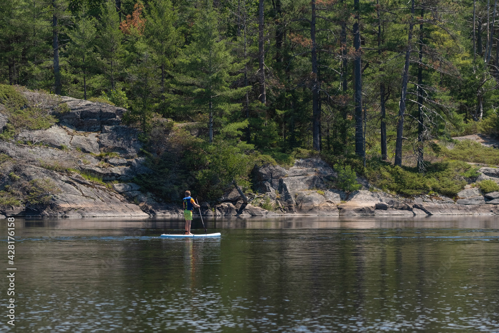 Young teenage boy in a life vest paddle boarding on a stand up board at Ontario Provincial Park. Summer recreational activities and water sports. Space for copy. Selective focus. Young teenage boy in a life vest paddle boarding on a stand up board at Ontario Provincial Park. Summer recreational activities and water sports. Space for copy. Selective focus.