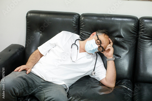 Medico in camice bianco e mascherina chirurgica , distrutto dalla fatica si accascia in un divano per riposarsi, 