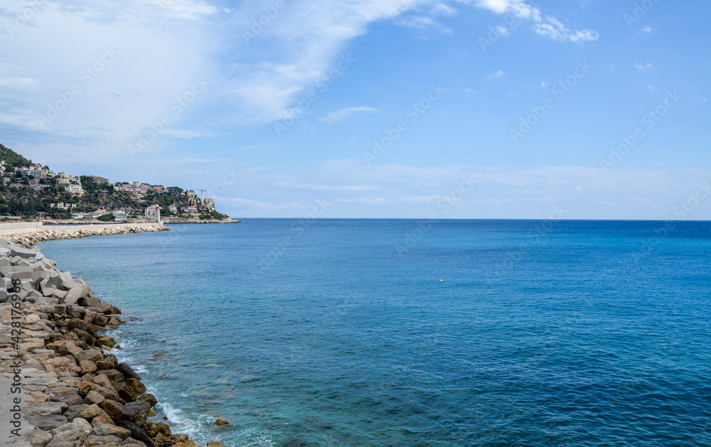 Stony seashore with beautiful blue sea water next to the lighthouse, visible in the background, Mediterranean sea, Nice, France