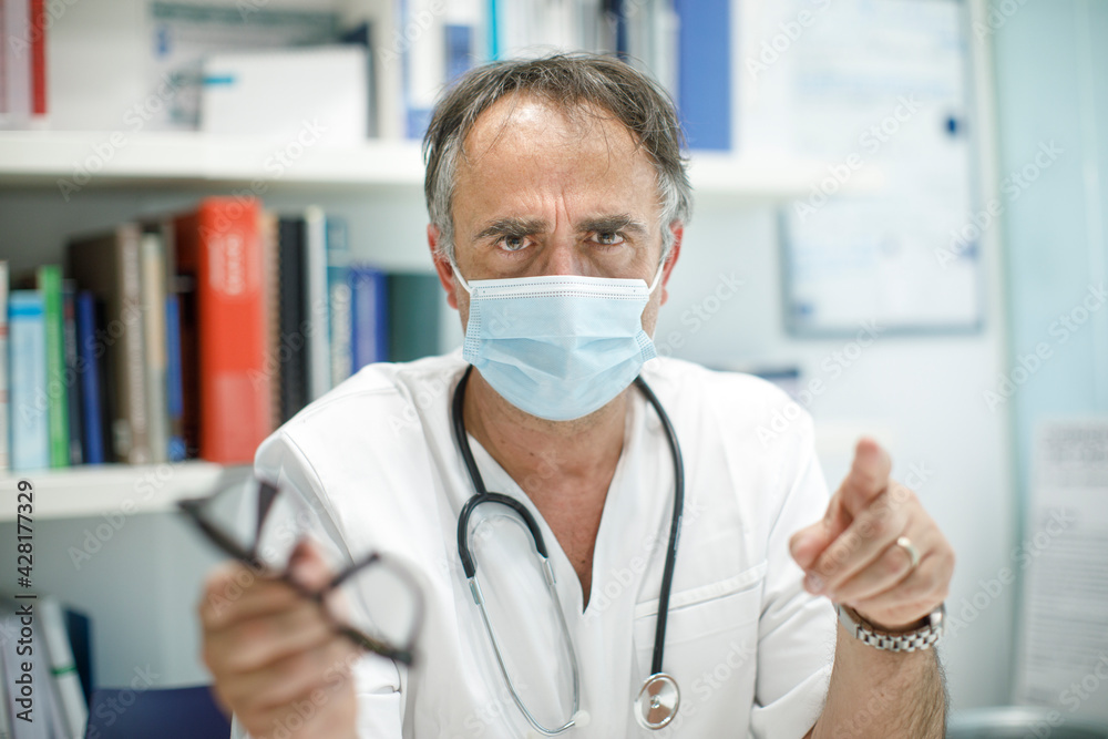 Medico cinquantenne in camice bianco e mascherina protettiva, seduto nella scrivania del suo ufficio,  guarda co espressione severa.