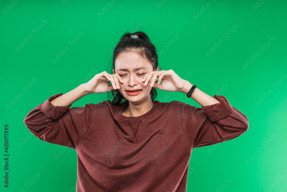 crying expression of a young Asian woman while wiping the tears with both hands isolated on green background