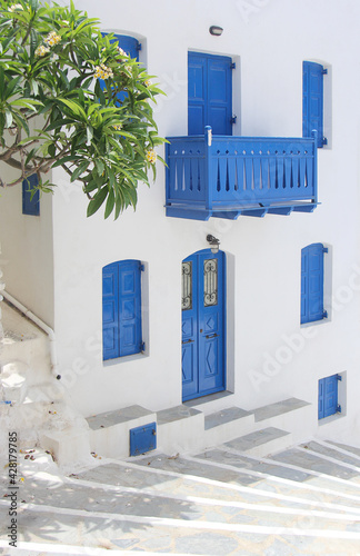 Nice and cozy courtyard in the Greek style. Windows with wooden blue shutters, plumeria blooms next to the porch. Summer sunny day on a Greek island. photo