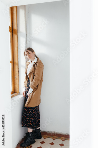 Stylish young girl stands on the background of a white wall near the window and looks at the camera