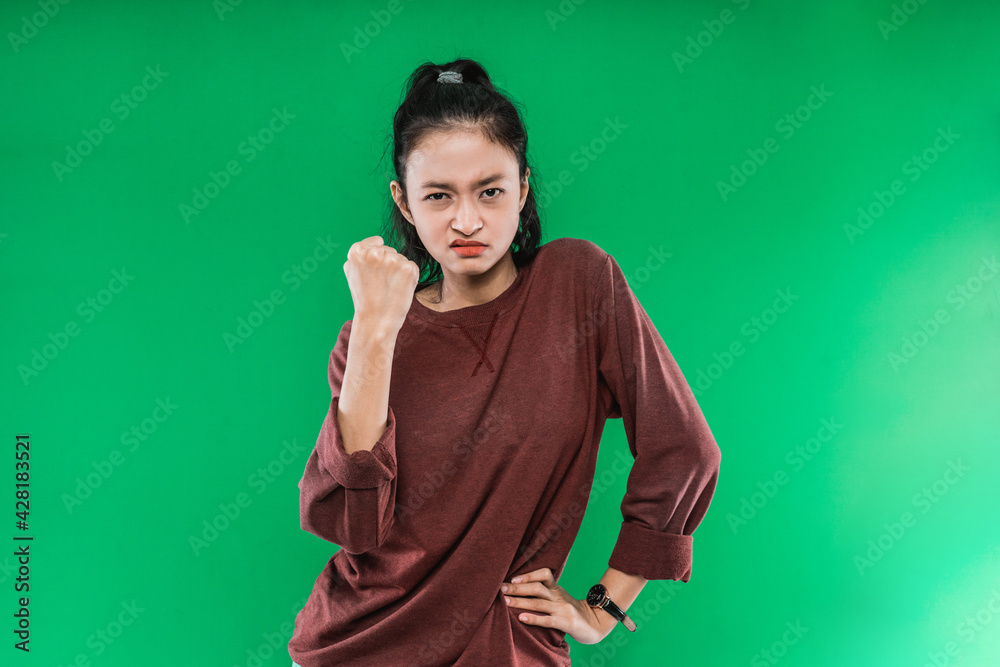 young woman face with an angry expression while clenching one fist and one hand on her hips against a green background young woman face with an angry expression while clenching one fist and one hand on her hips against a green background